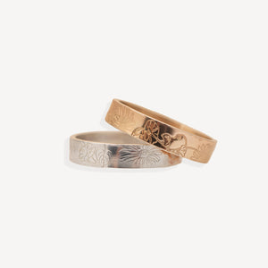 Two engraved metal bands, one rose gold and one silver, overlapping on a clean off-white background. Both bands feature a floral pattern engraved on the outer surface.