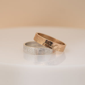Two engraved rings, one silver-toned and one rose gold-toned, displayed on a white reflective surface against a soft beige background.