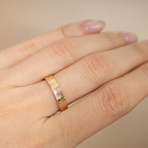 A close-up of a hand wearing a thin rose gold band ring with a sunburst engraved pattern, set against a soft beige background.