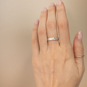 A close-up of a hand wearing a textured silver band ring on the ring finger, with a thin gold band ring on the middle finger, set against a solid beige background.