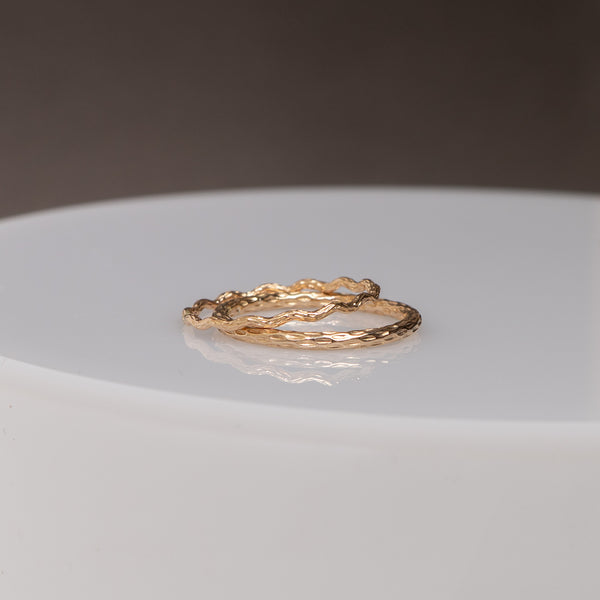 Two textured gold rings with organic, wavy shapes resting on a white circular pedestal against a dark gray background.