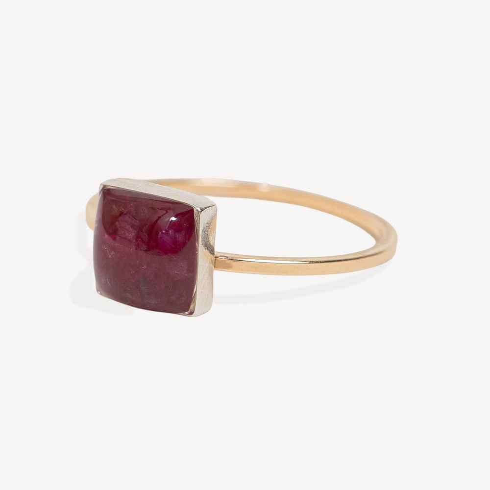 A square-cut deep red ruby gemstone ring set in a polished gold band, photographed on a clean, light off-white background.