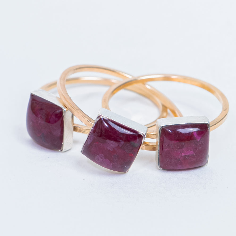 A square-cut deep red ruby gemstone ring set in a polished gold band, photographed on a clean, light off-white background.