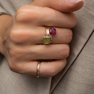 A close-up of a hand wearing a gold ring with two square gemstones, one green and one pinkish-red, resting on beige fabric.