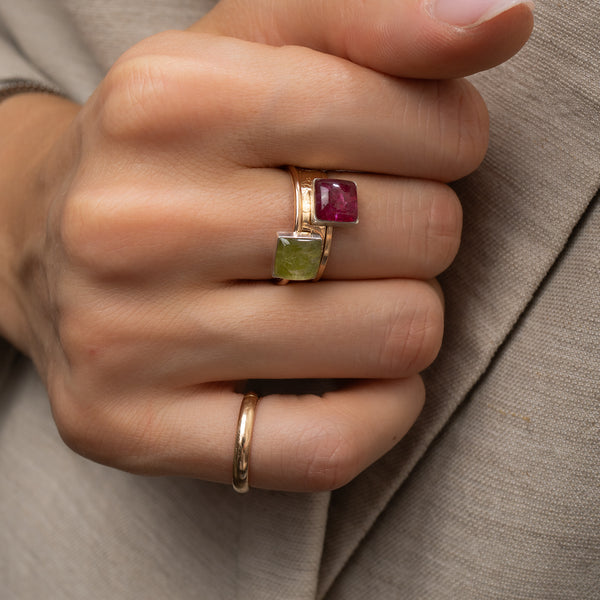 A close-up of a hand wearing a gold ring with two square gemstones, one green and one pinkish-red, resting on beige fabric.