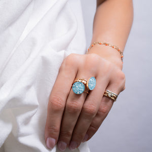 A close-up of a hand wearing two gold rings with oval light blue stones, along with a delicate gold and white beaded bracelet, set against a soft white draped fabric background.