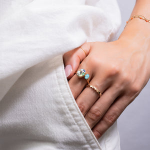 A close-up of a person's hand wearing a gold ring with a large round clear gemstone and a small light blue stone, along with a delicate gold beaded ring and a gold bracelet, styled with a white top.