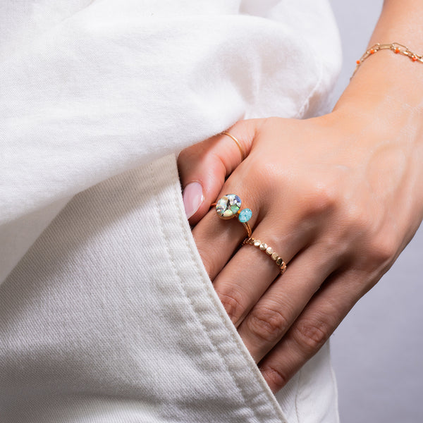 A close-up of a person's hand wearing a gold ring with a large round clear gemstone and a small light blue stone, along with a delicate gold beaded ring and a gold bracelet, styled with a white top.