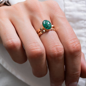 A close-up of a hand wearing a rose gold ring with a round green emerald-cut gemstone, set against a soft white background.