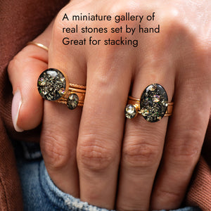 A close-up of a hand wearing multiple gold rings with dark, glittery real stones, overlaid with the text 'A miniature gallery of real stones set by hand Great for stacking'.