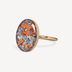 An oval ring featuring a textured, metallic resin cabochon with orange, silver, and blue flecks, set in a polished gold-tone band against a light gray background.