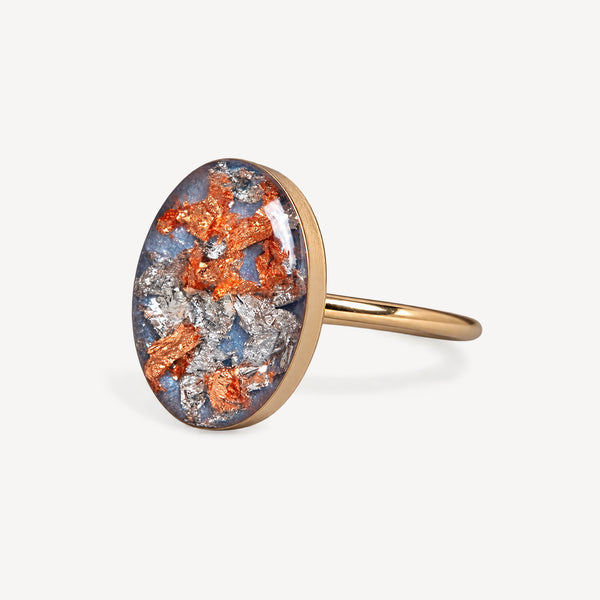 An oval ring featuring a textured, metallic resin cabochon with orange, silver, and blue flecks, set in a polished gold-tone band against a light gray background.