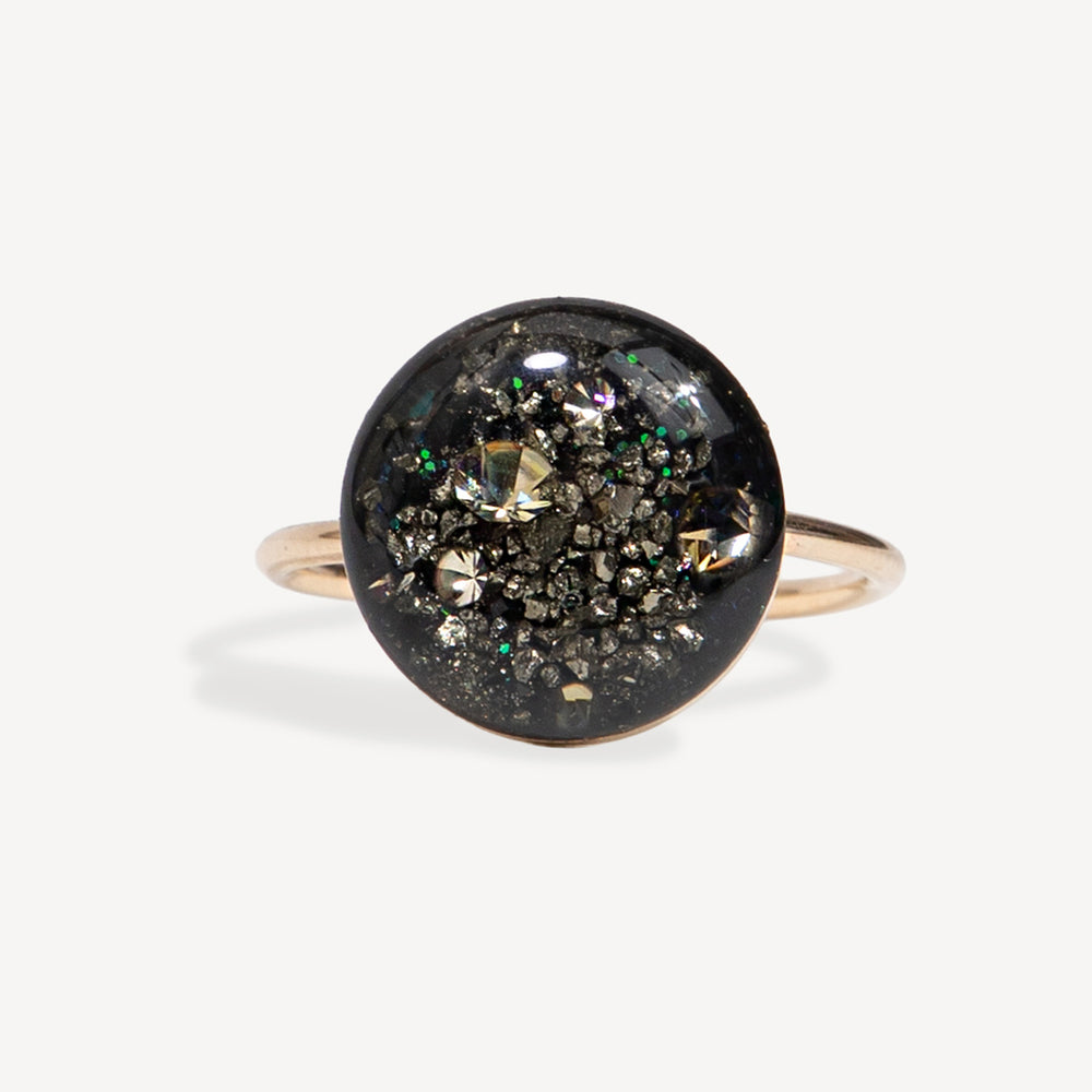 A round bezel-set ring featuring a black background filled with metallic gold, silver, and iridescent glitter, mounted on a slim rose gold band.