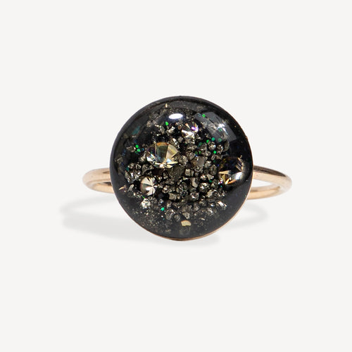 A round bezel-set ring featuring a black background filled with metallic gold, silver, and iridescent glitter, mounted on a slim rose gold band.