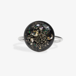 A silver band ring featuring a large, round, domed black resin cabochon filled with metallic gold and silver glitter, set against a clean white background.