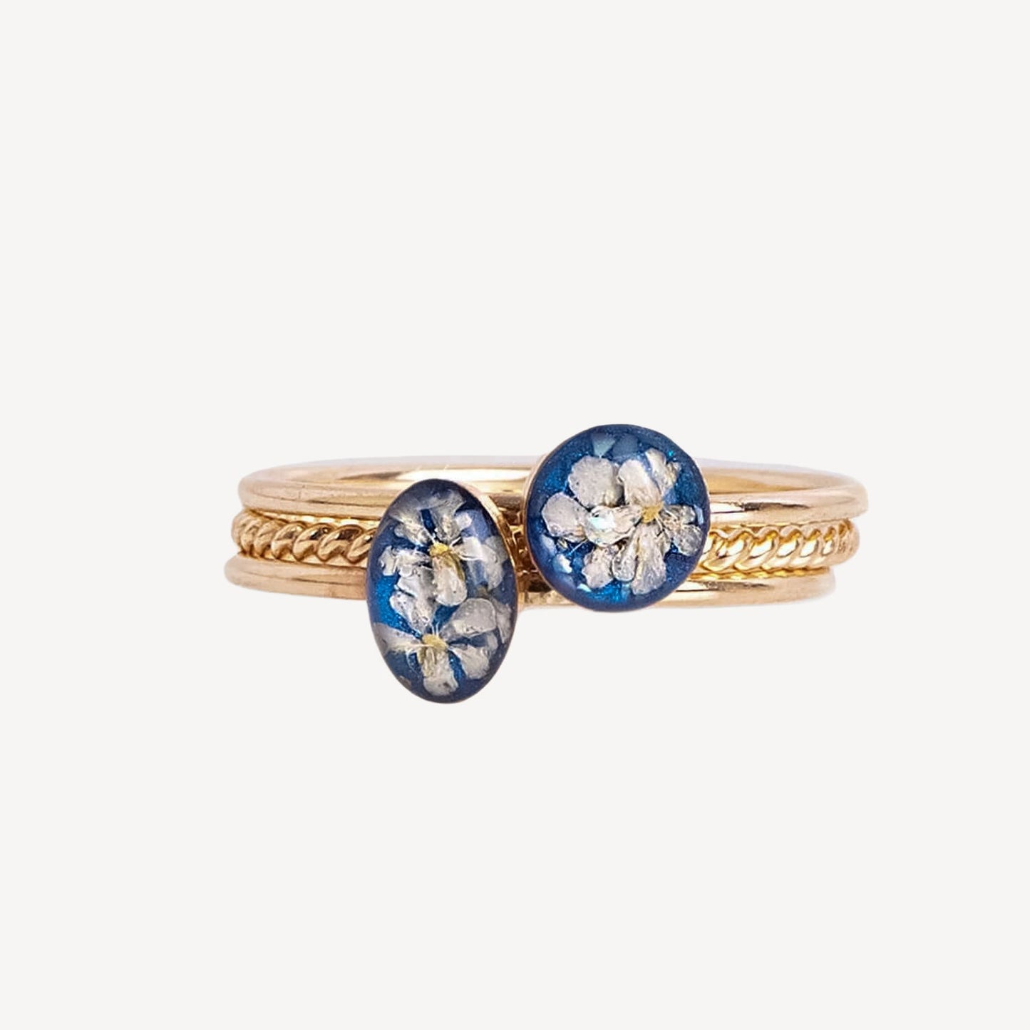 A gold ring stack featuring two oval blue resin cabochons with pressed white flowers, set against a plain light gray background.
