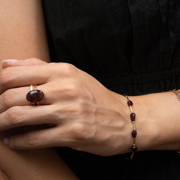 A close-up lifestyle shot of a person's hand and wrist wearing jewelry. The hand wears a gold ring with a large oval dark red  Garnet gemstone, and the wrist wears a delicate gold chain bracelet with matching dark red oval Garnet beads.