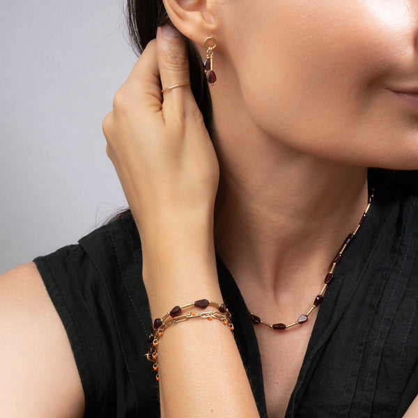 A close-up lifestyle shot of a woman wearing a matching jewelry set, featuring a gold chain necklace with dark brown Garnet gemstone beads, a matching bracelet, and a small gold hoop earring.