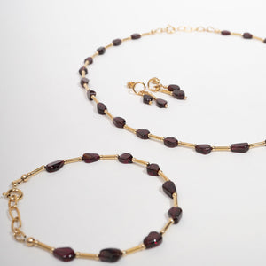 Gold-tone jewelry set featuring a Garnet beaded necklace, bracelet, and matching drop earrings with dark reddish-brown faceted beads on a white background.