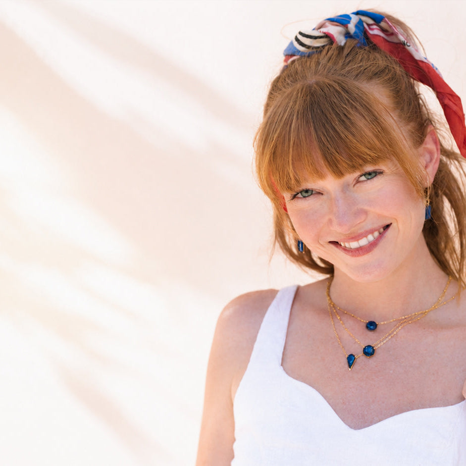 A smiling woman with red hair tied up with a red, blue, and black patterned scarf, wearing a white top and a gold necklace with dark blue beads, set against a bright, sunlit beige background with soft pink floral shadows.