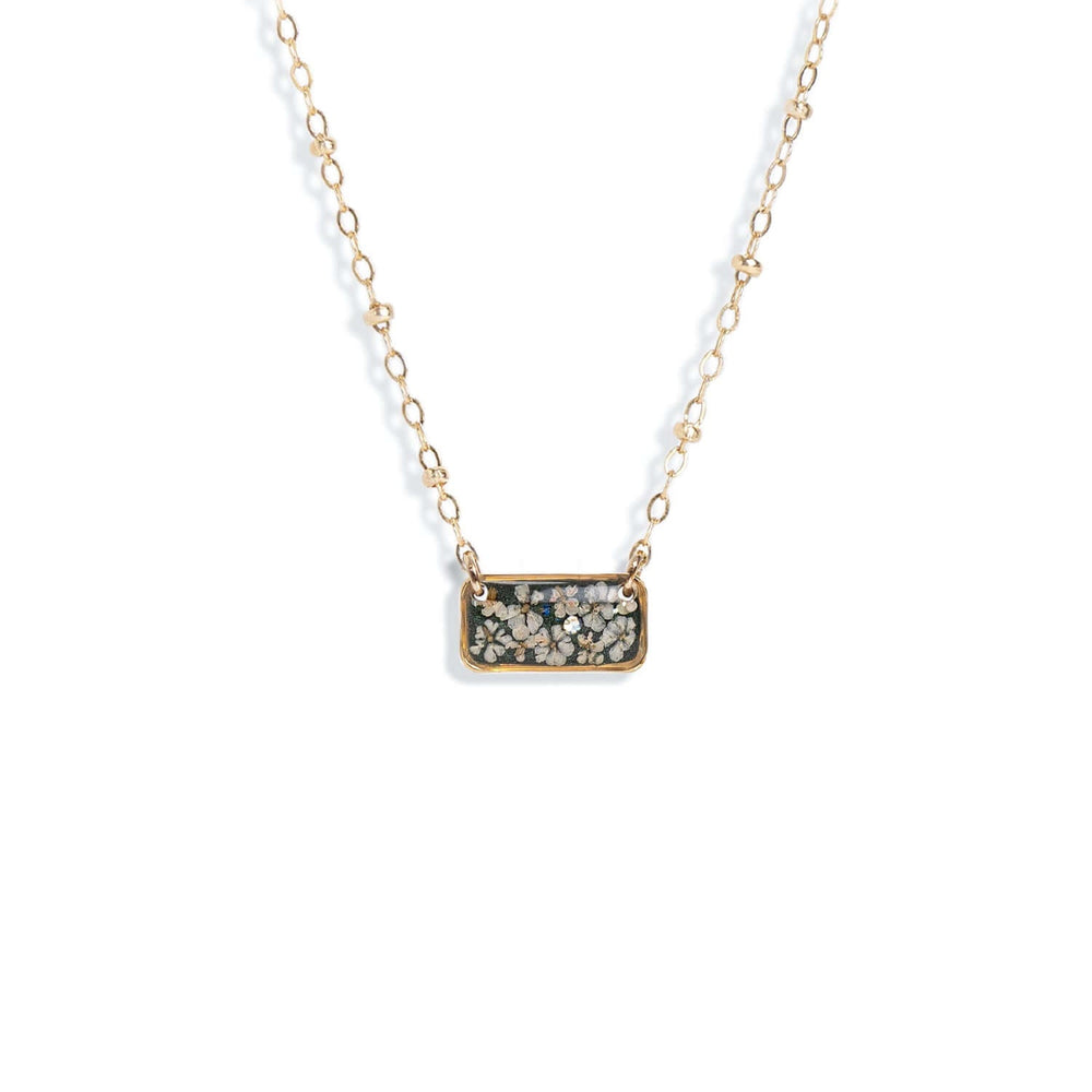 A gold chain necklace featuring a rectangular pendant filled with dried flowers and small botanical pieces, shown against a plain off-white background.
