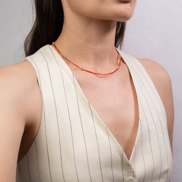 Woman wearing a white sleeveless top with a red beaded necklace against a gray background