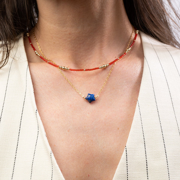 Close-up of a person wearing two necklaces, one red and one gold with a blue pendant, on a white background.