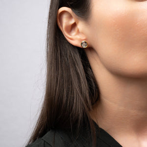 Close-up of an ear wearing a gold earring with a clear gemstone on a neutral background