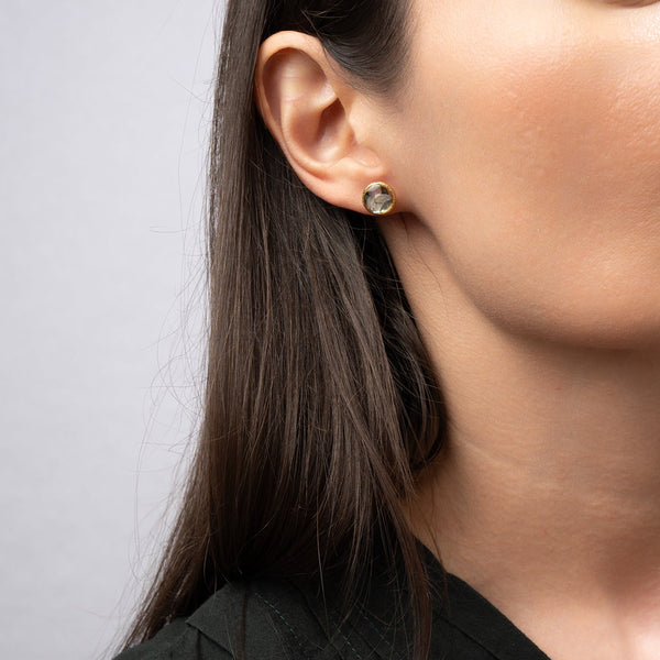 Close-up of an ear wearing a gold earring with a clear gemstone on a neutral background