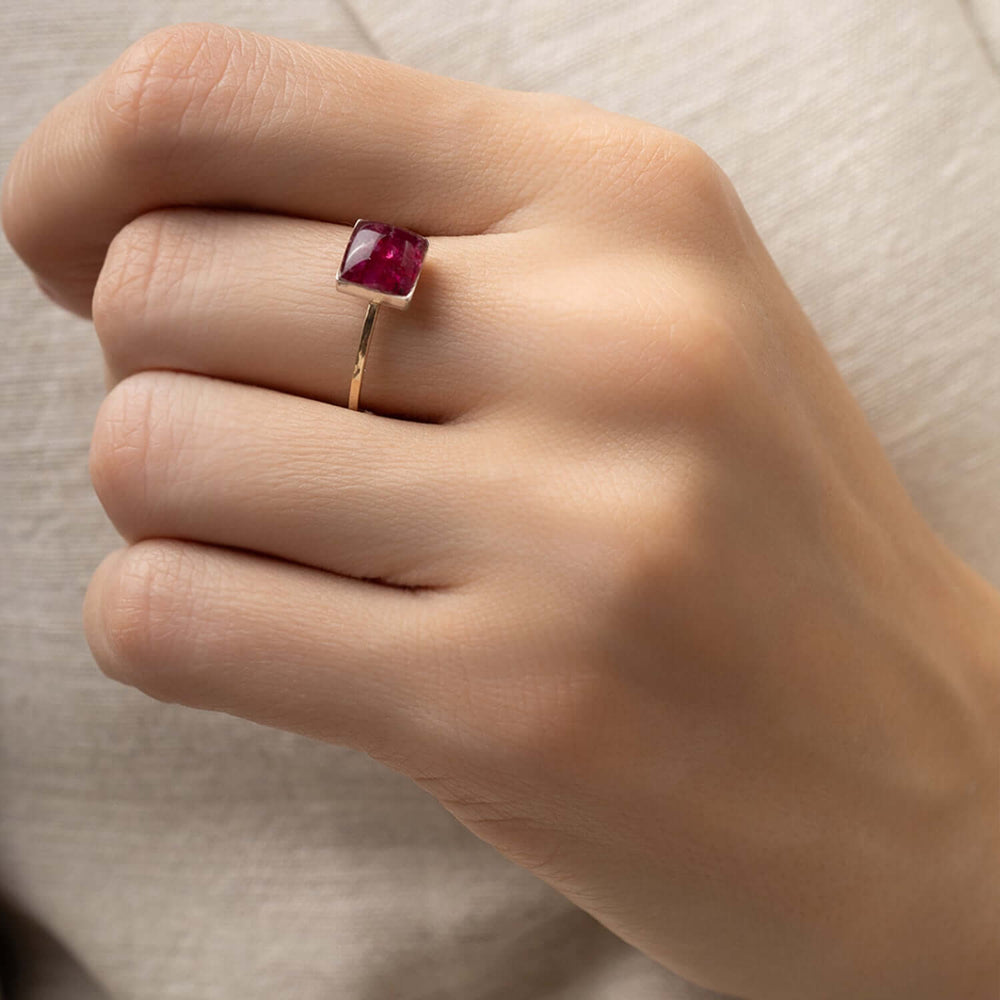 A square-cut deep red ruby gemstone ring set in a polished gold band, photographed on a clean, light off-white background.