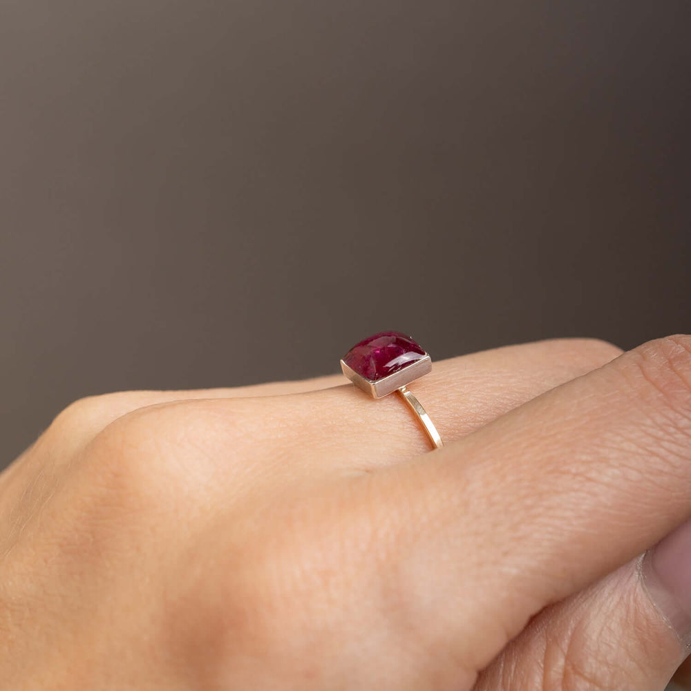 A square-cut deep red ruby gemstone ring set in a polished gold band, photographed on a clean, light off-white background.