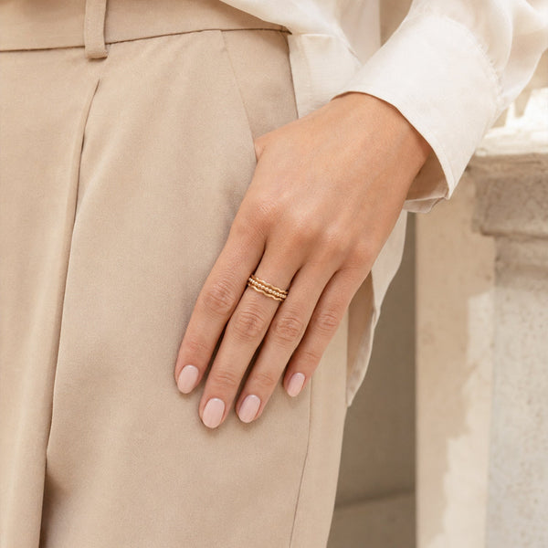 A close-up of a person's hand wearing a delicate gold ring, resting on their lap. The person is wearing beige trousers and a white blouse, with a soft, blurred stone wall in the background.