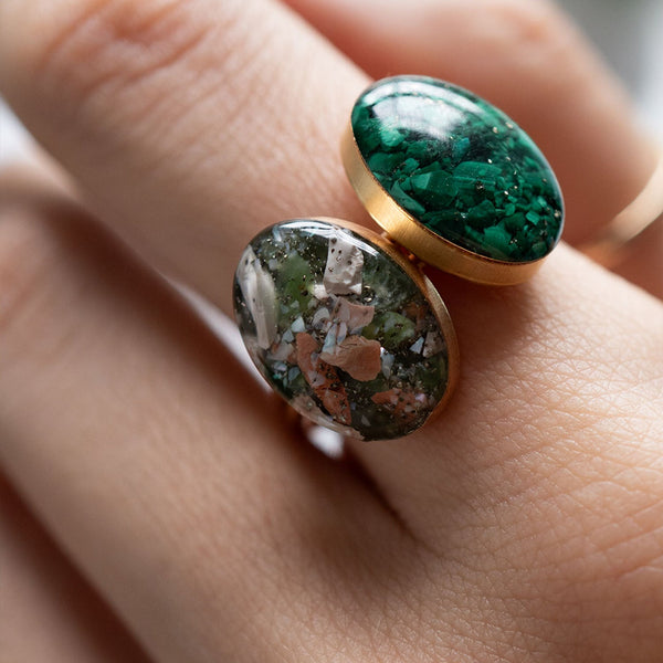 A close-up of a gold ring worn on a finger, featuring two oval green stone cabochons with different marbled patterns.