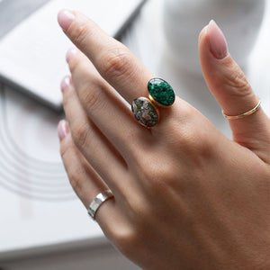 A close-up of a hand wearing two rings: a larger gold ring with oval green and brown marbled stones, and a smaller gold band ring on another finger, against a soft white background.