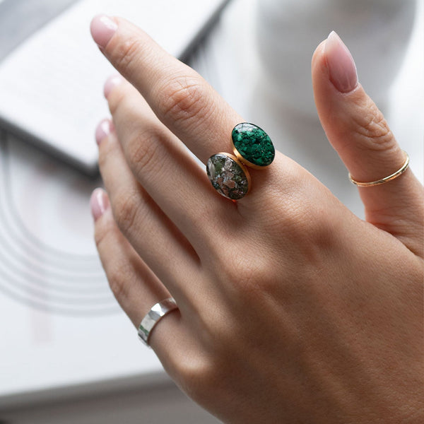 A close-up of a hand wearing two rings: a larger gold ring with oval green and brown marbled stones, and a smaller gold band ring on another finger, against a soft white background.