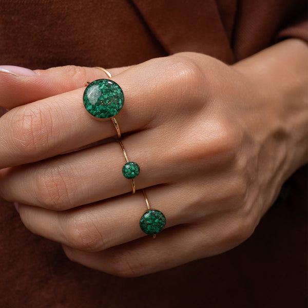 A close-up of a hand wearing a three-stone gold ring set with round green marbled gemstones, set against a dark brown background.