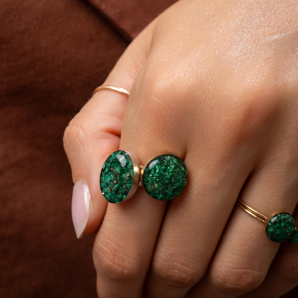 A close-up of a hand wearing three gold rings, with two rings featuring oval green glittery stones. The background is a soft, textured brown fabric.
