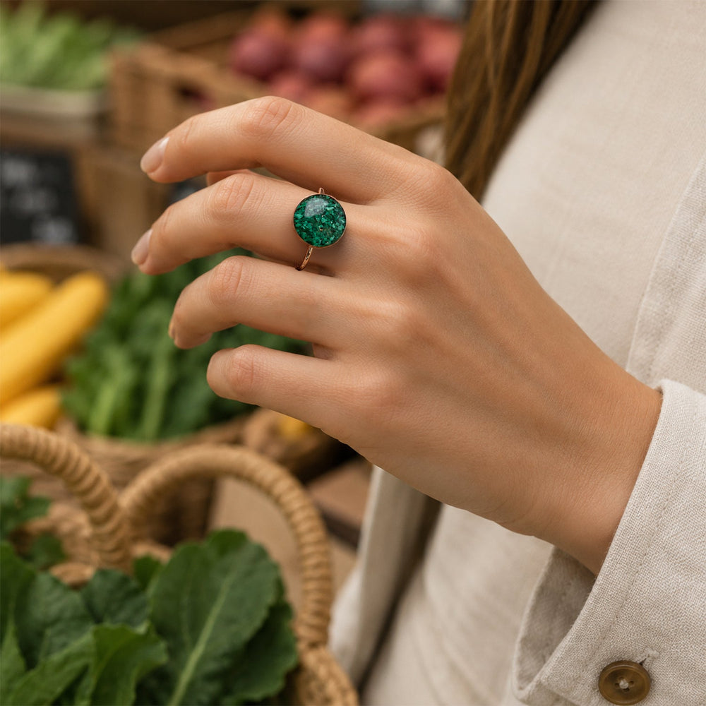 A gold band ring featuring a large, round cabochon stone filled with sparkling green glitter, set against a clean white background.