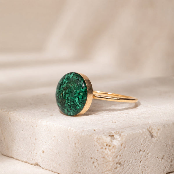 A gold ring with a round green malachite stone, resting on a textured beige stone surface.