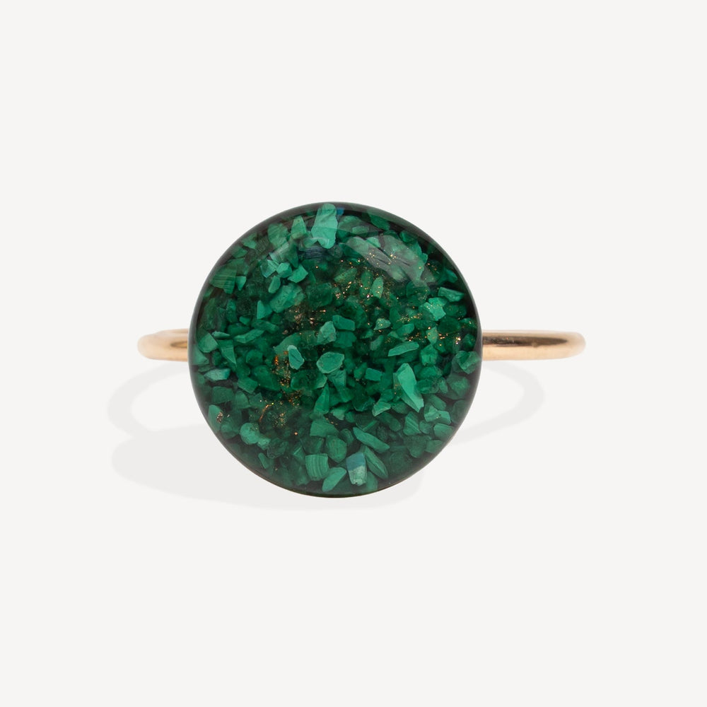 A gold band ring featuring a large, round cabochon stone filled with sparkling green glitter, set against a clean white background.