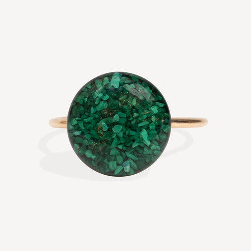 A gold band ring featuring a large, round cabochon stone filled with sparkling green glitter, set against a clean white background.