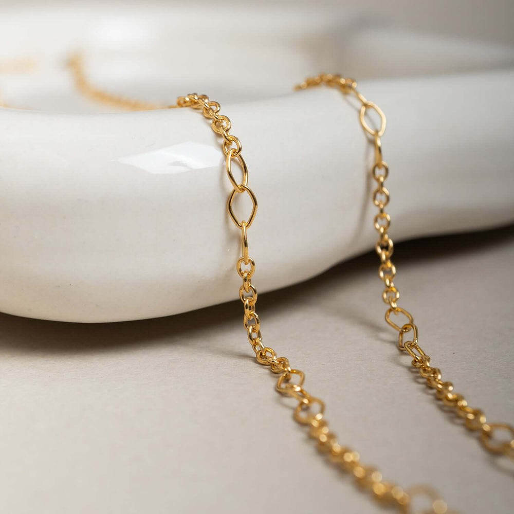 A delicate gold chain necklace with elongated oval links, displayed in a curved shape against a clean, off-white background.