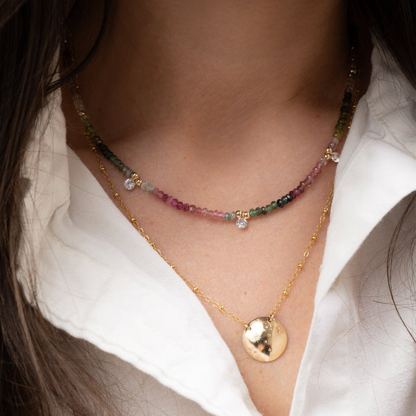 Close-up of a person wearing two necklaces, one with Watermelon Tourmaline Beaded Necklace and the other with a gold round pendant, on a white shirt.