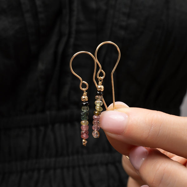 Gold earrings with multicolored beads held by a hand against a black background